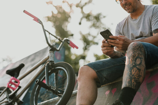 Cropped picture of a middle-aged tattooed urban man sitting in a skate park and reading messages on the phone.