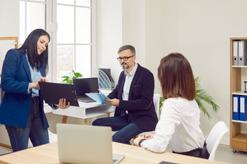 Young woman employee showing something to her colleagues on the workplace in the office. Three coworkers discussing a new projects or startups, analyzing company. Team work concept.