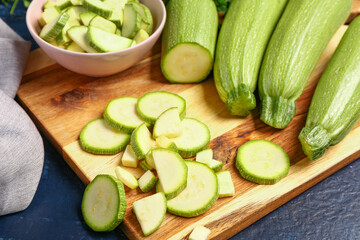 Board with fresh green zucchini and bowl of slices on blue background