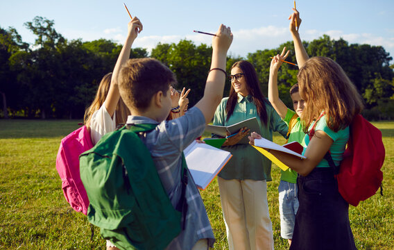 Group of school children learning about nature during outdoor lesson. Smiling teacher asks questions about environment. Students standing on green grass meadow, holding notebooks and raising hands up