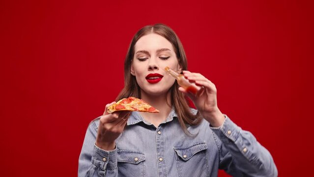 Beautiful yougn girl with red lips emotional eating slice of pizza with hot chilli sauce. Model posung agasint red backgorund. Burning mouth, spicy taste. Concept of food, taste, human emotions. Ad