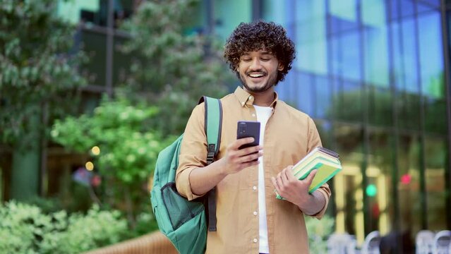 Handsome smiling university student with backpack and books in hands using smartphone standing in campus. Male chats, corresponds online with friends, browses social networks, checks email messages