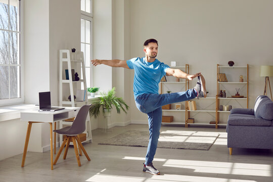 Man doing a leg swing during a fitness workout at home. Fit, sporty, handsome young man in a blue T shirt and jeans doing leg swing exercise in a modern sunny living room interior
