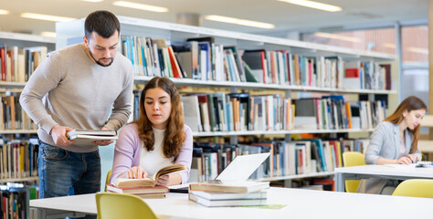 Guy and a girl students studying at the university, preparing for the exam in the library studying on a laptop and looking ..for sources of information in books