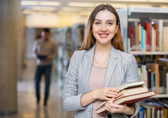 Positive female librarian standing near bookshelves with book in hands