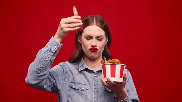 Funny crazy face. Emotional young girls eating fried chicken with hot chilli sauce against red studio background. Burning mouth. Spicy taste. Fast food lover. Concept of food, taste. Ad