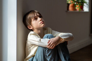 Lonely little boy sadly looking up hugging knees sits near window at home alone. Upset unhappy...