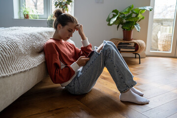 Teens and cyberbullying. Upset teen girl sitting on floor near bed using smartphone at home, scrolling social media. Child spending too much time on phone. Teenagers and gadget addiction - obrazy, fototapety, plakaty