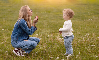 Young mother plays with her little son in the park. Dandelions pick flowers and blow
