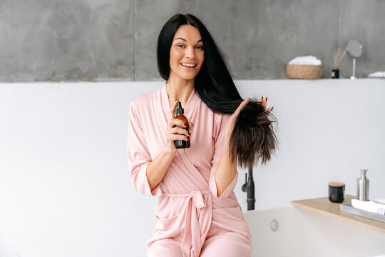 Female beauty routine. Happy brunette caucasian woman applying essential oil spray, fluid, on her long hair, sits in a bathroom, looks at camera, smiles. Haircare concept. Split ends repair treatment