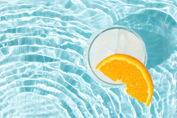 Glass of ice cubes with slice of fresh orange in rippled water of pool. Summer cold drink...