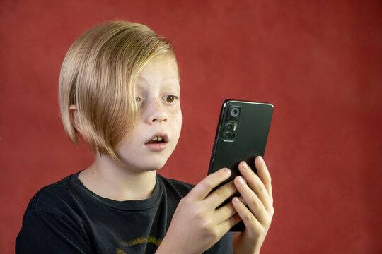 Niño rubio asombrado con teléfono movil celular fondo rojo
