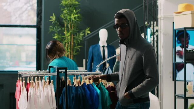 African american thief robbing clothing store, analyzing racks with stylish clothes. Robber trying to steal fashionable merchandise, wearing sunglasses and hood not to be recognized