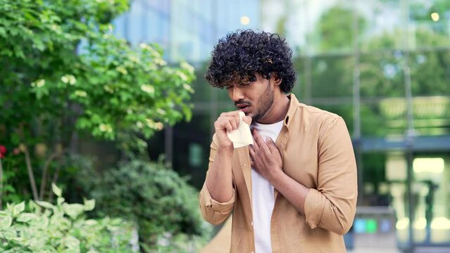 Young man coughs suffering from a cold while standing near an office building. Sad male sneezes into a handkerchief, holds his hand on his chest. He has flu, bronchitis or respiratory tract infection