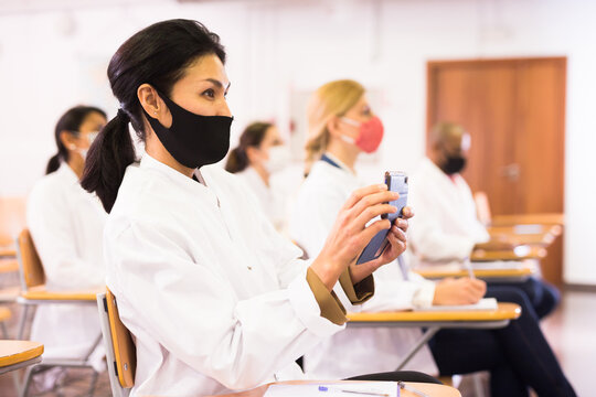Confident woman participant of training program for health workers recording lecture on smartphone at conference hall, all people wearing face masks for viral protection
