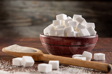 White sugar cubes in wooden bowl and granulated sugar on wooden spoon.