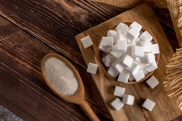 White sugar cubes in wooden bowl and granulated sugar on wooden spoon.