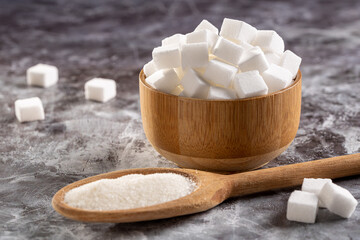 White sugar cubes in wooden bowl and granulated sugar on wooden spoon.