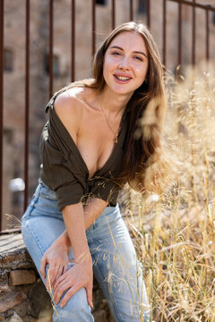 Portrait of charming young adult woman with long hair wearing jeans and unbuttoned khaki blouse standing in sensual pose near rusted fence against old abandoned building, having positive expression