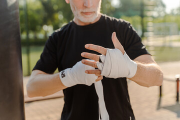 Details on the hands of senior male boxer tying elastic bandage on his wrists before boxing outdoors