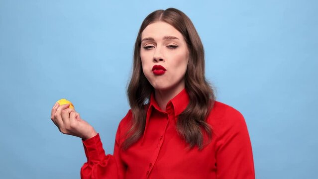 Young beautiful girl biting lemon against blue studio background. Emotional woman with grimacing face eating sour citrus. Concept of taste, human emotions, facial expression