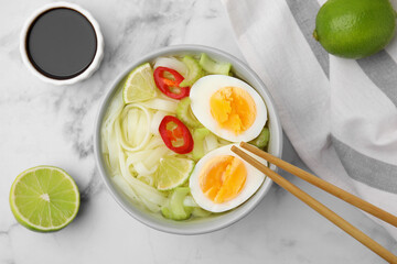 Bowl of delicious rice noodle soup with celery and egg on white marble table, flat lay