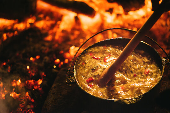 A wooden ladle stirs a pot of peppery soup in a cast iron pot over red hot coals