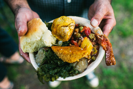 Hands hold a plate overloaded with food from a bbq including bacon, biscuits and greens in an outdoor setting