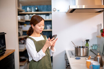 Fototapeta na wymiar キッチンでスマホを見る、エプロン姿の若い日本人の女性