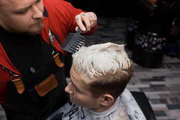 The hand of a professional hairdresser with a comb, combs the client's hair in white dye in the process of dyeing hair in a barbershop