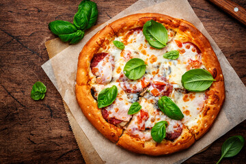 Pizza with spicy salami sausage, mozzarella cheese, tomato sauce and green basil on rustic wooden kitchen table background, top view