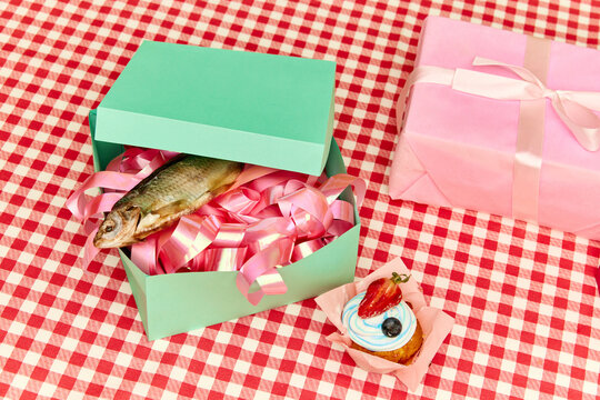 Extraordinary gift. Top view of present box with fish lying on checkered tablecloth with cakes. Layout. Concept of party, celebration, surprise. Crazy idas. Pop art
