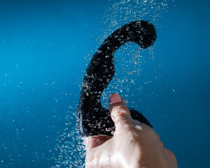 A woman washes a black prostate stimulator under running water on a blue background. Sex toy...
