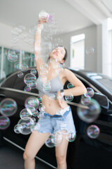 Sexy woman play bubble machine wearing grey bra and denim shorts pose with black car.