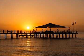 Fototapeta na wymiar Pier at dawn. Wooden pier on the azure sea at dawn in orange tones, wallpaper, seasonal background. Orange dawn on the sea, Beldibi, Antalya, Türkiye.