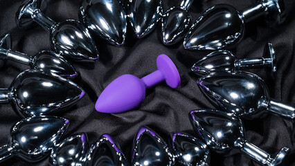 Purple silicone butt plug among many metal ones on a black silk sheet. 
