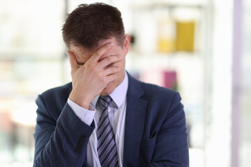Tired employee in formal suit covers face with hand in room