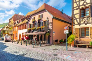 A picturesque cobbled street of shops and restaurants in the historic medieval Alsatian village of Kaysersberg-Vignoble France, near Colmar along the wine route. - obrazy, fototapety, plakaty