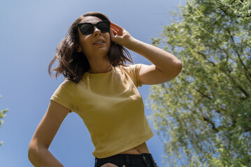 Bottom view of happy millennial woman in yellow t-shirt and sunglasses smiling broadly, looking directly to the camera.