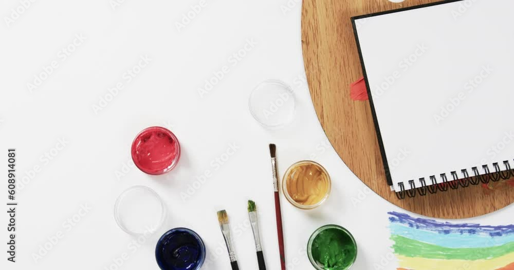 Poster overhead view of notebook, brushes and paints on white background with copy space, in slow motion