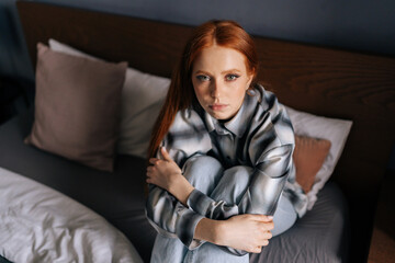 High-angle view portrait of sad young woman hugging knees sitting on bed, feeling lonely goes...