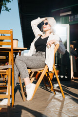 Stylish young woman in sunglasses posing at street cafe table on sunny day. Charming, glamorous caucasian blonde model chilling with cup of coffee outdoors, looking away