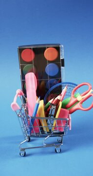 Vertical video of close up of shopping trolley with school items and paints on blue background