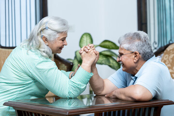 indian senior couple doing arm wrestling at home in living room , Retired life fun concept shoot