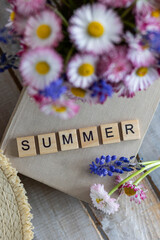 Colourful bouquet with blue flowers, meadow daisy in a cup on the table. Old book, straw hat, countryside home decor. Postcard for the holidays. Word summer written in wooden letters. Calendar