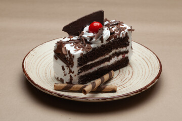 A piece of Delicious Chocolate pastry with a cherry and chocolate wafer on top. On a white plate. Front view.