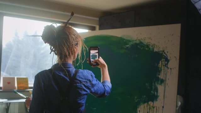 A talented red-haired female artist with dreadlocks takes a photo of a finished masterpiece on a smartphone in a loft studio. Creating an abstract painting for the Internet and an online exhibition.