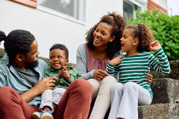 Happy black family talks while relaxing on staircase in front of their house.