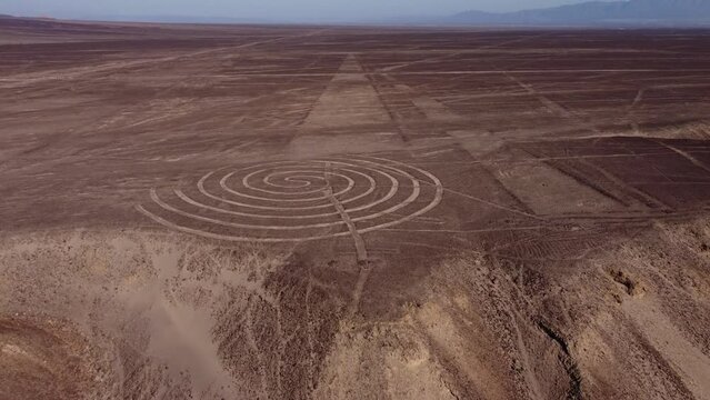 Spiral and long linear ancient geoglyphs drawn on flat desert plateau. Part of the world famous Nazca Lines. Drone flies forward tilting camera up to the horizon. Located in Nazca, Peru.