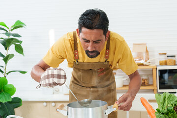 Bearded Asian man in kitchen, wearing heat resistant gloves Use a ladle to stir the hot soup in the...